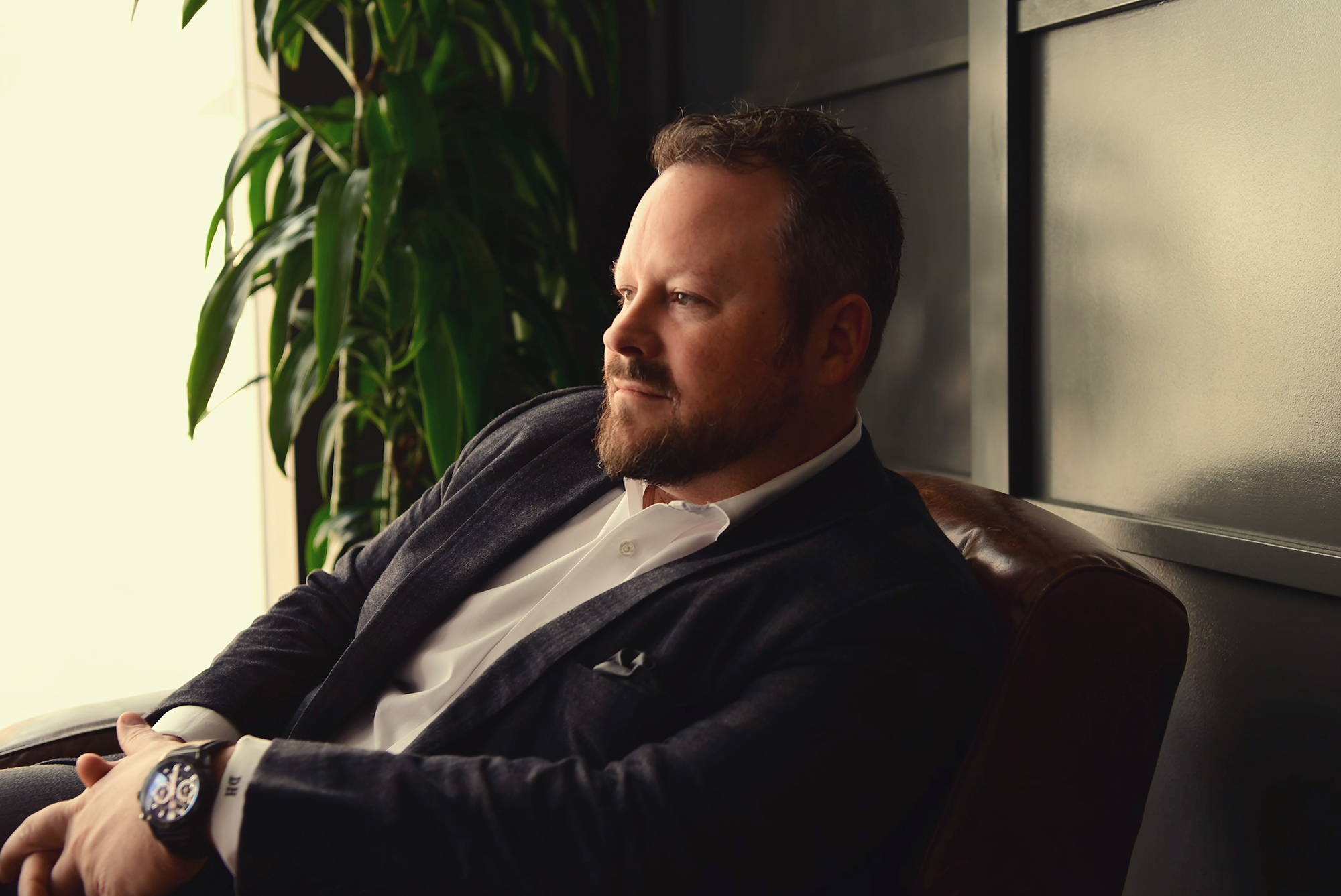 A man with a beard sits on a leather chair, wearing a suit jacket and shirt. He looks to the side, with a plant in the background.