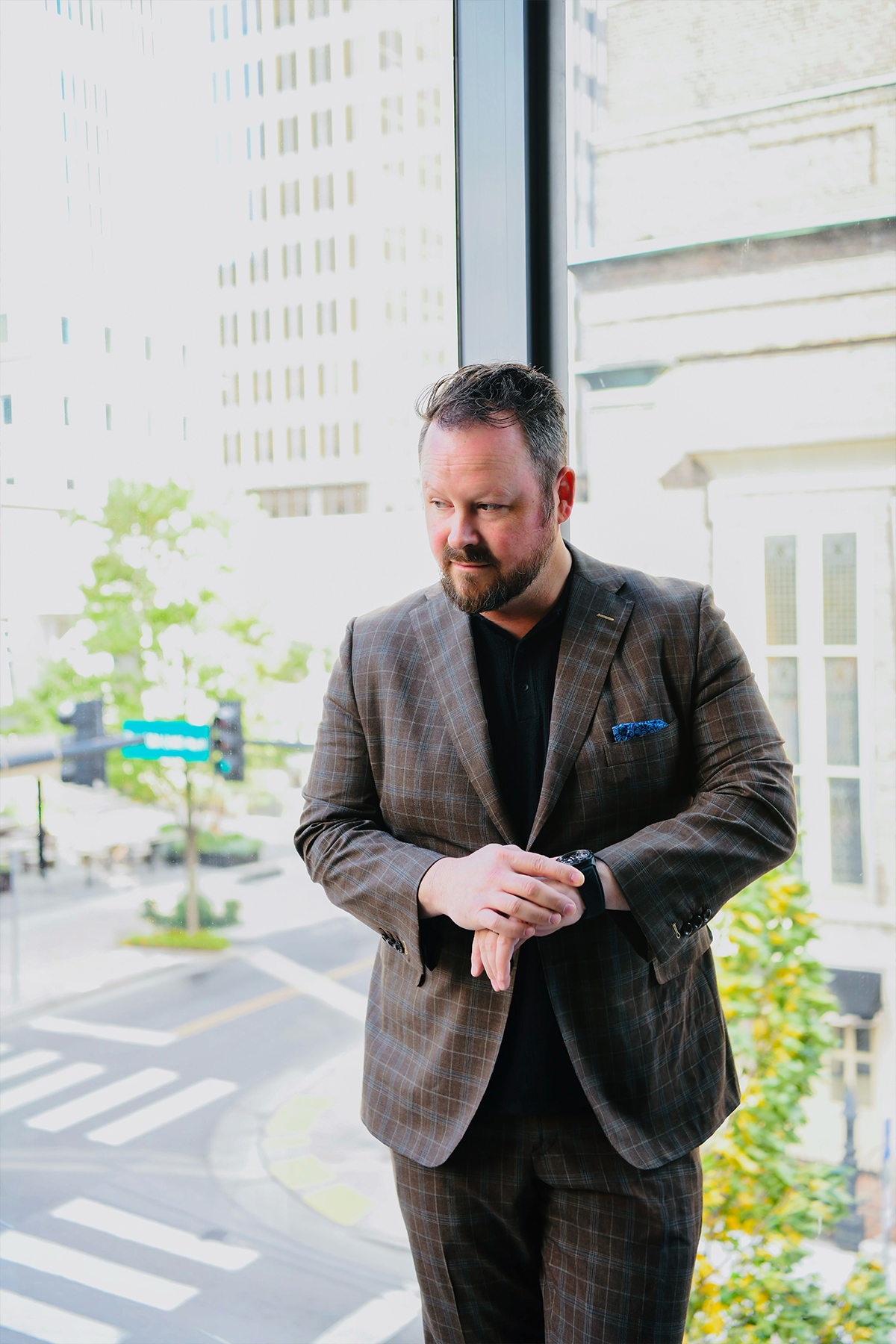 A person in a plaid suit stands by a window, looking down. Urban buildings and streets are visible outside.