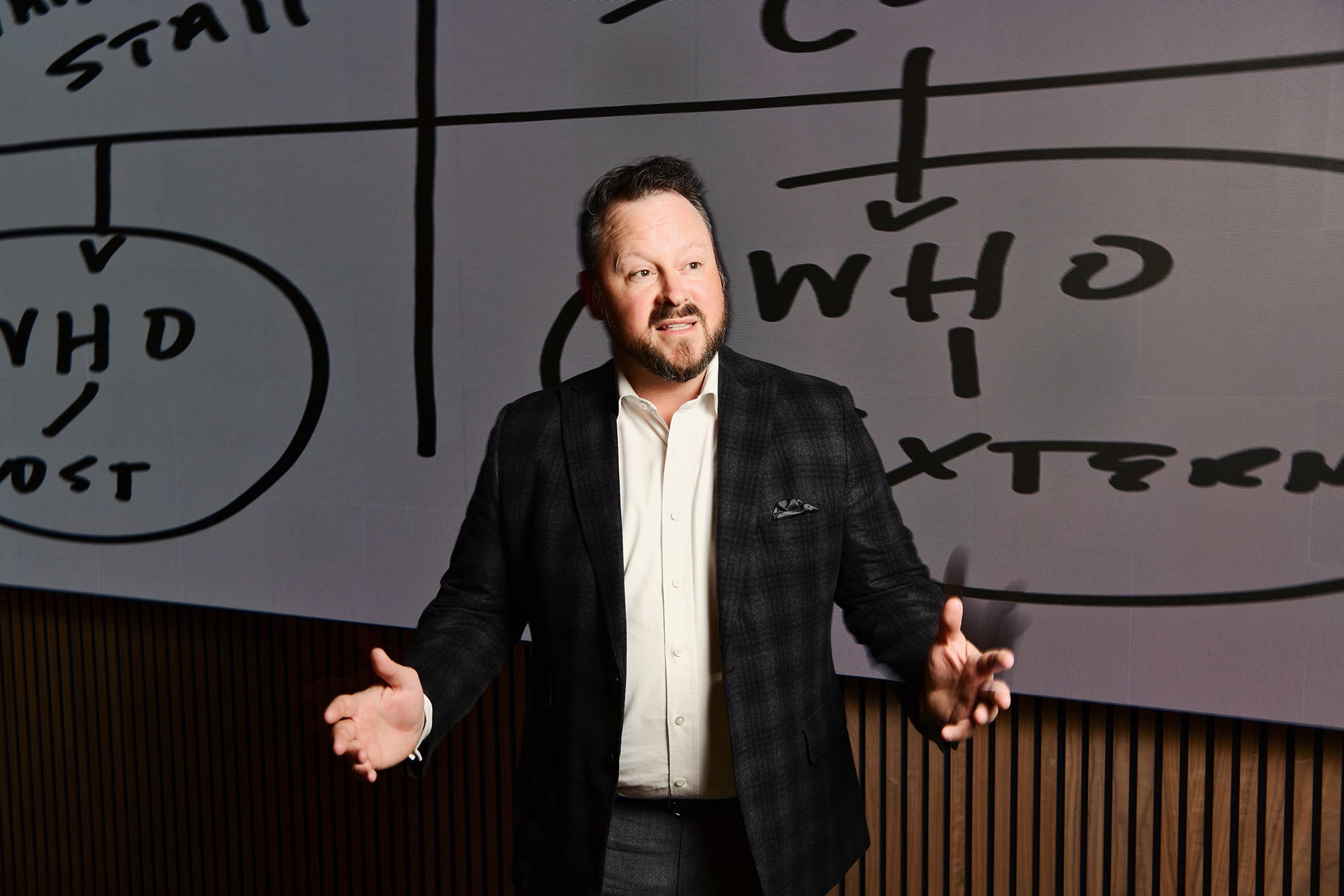 A man in a suit stands in front of a screen with a diagram, gesturing with his hands.