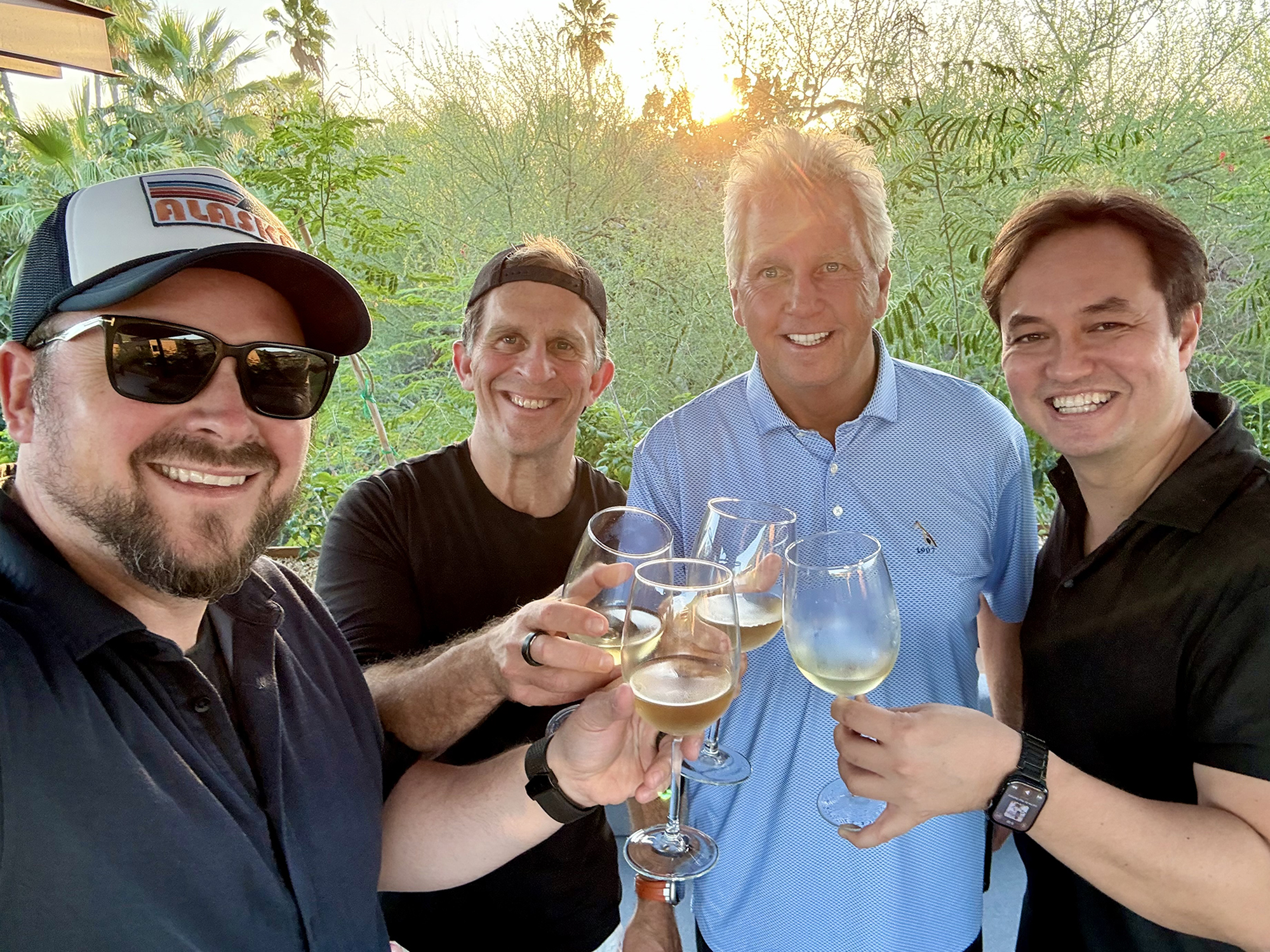 Four men smiling and holding wine glasses outdoors with greenery in the background.