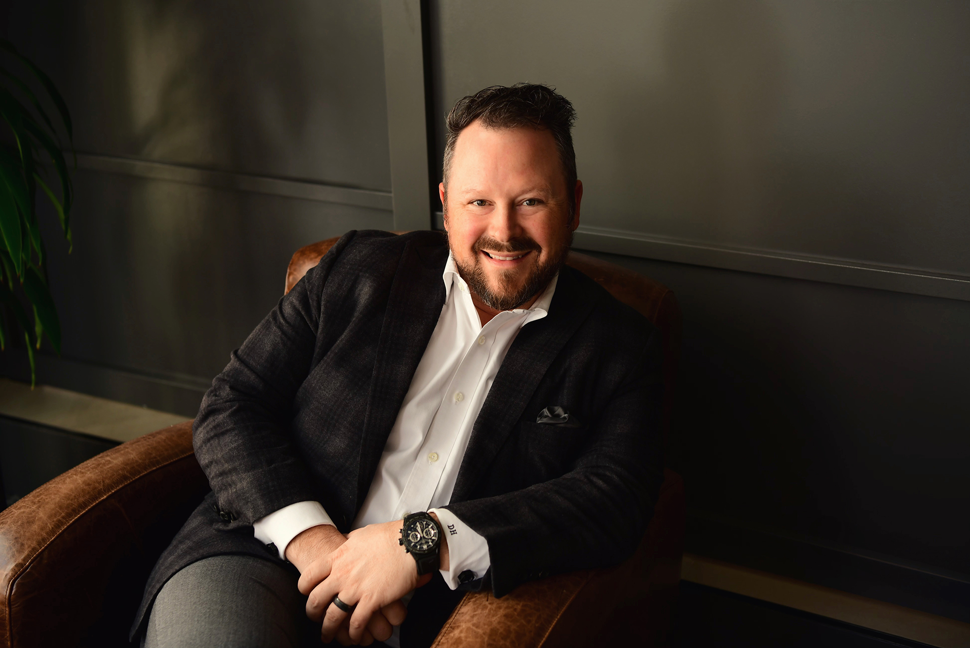 A man in a dark blazer and white shirt sits in a brown leather chair, smiling at the camera.