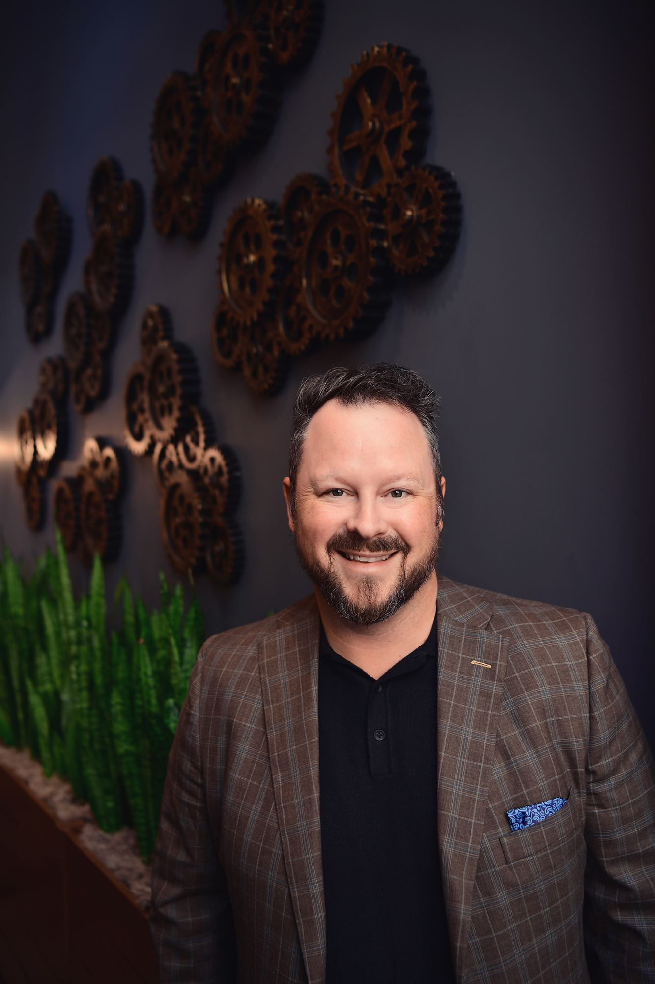 A man with a beard and a plaid blazer stands in front of a dark wall decorated with metal gears.