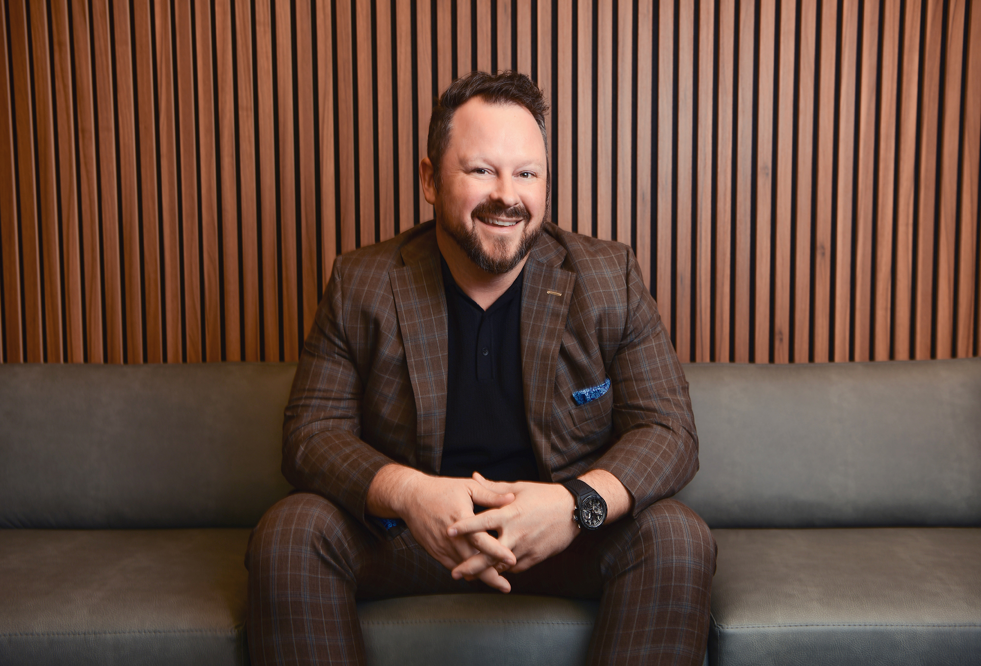 A person in a brown plaid suit sits on a gray couch, smiling. The background features vertical wooden slats.