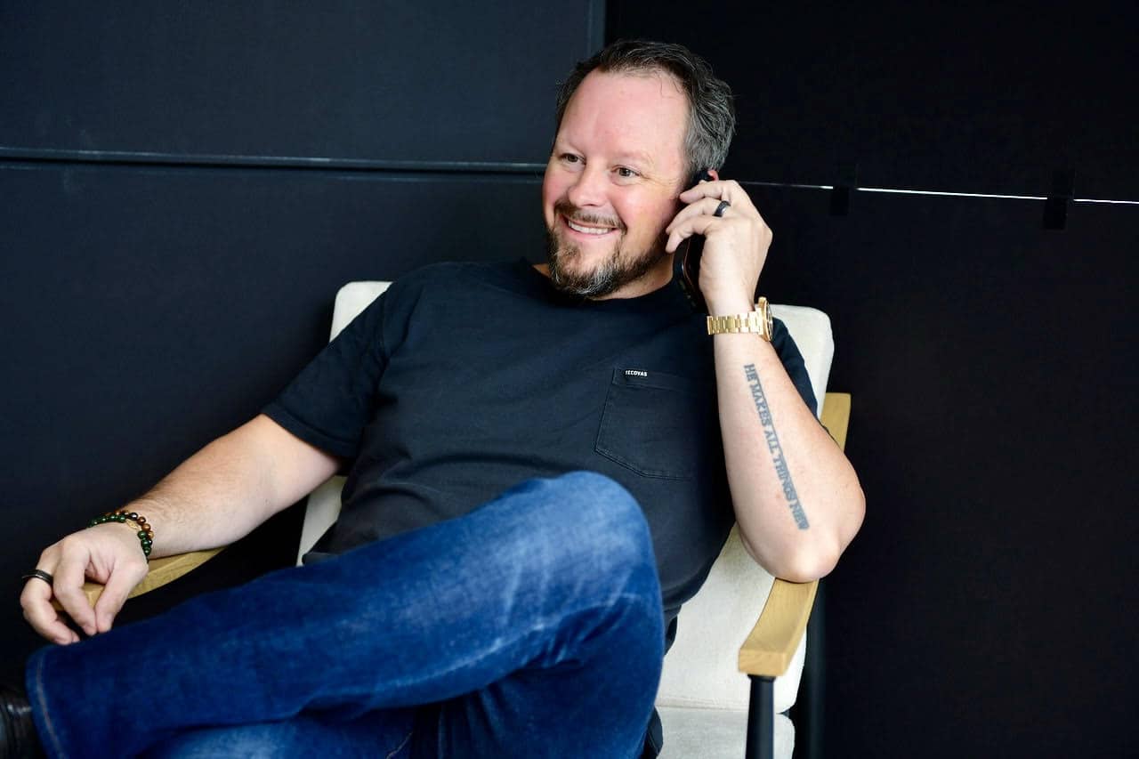 A man sits in a chair, smiling and talking on a phone. He is wearing a black t-shirt, blue jeans, and a gold watch, with one arm resting on the armrest.