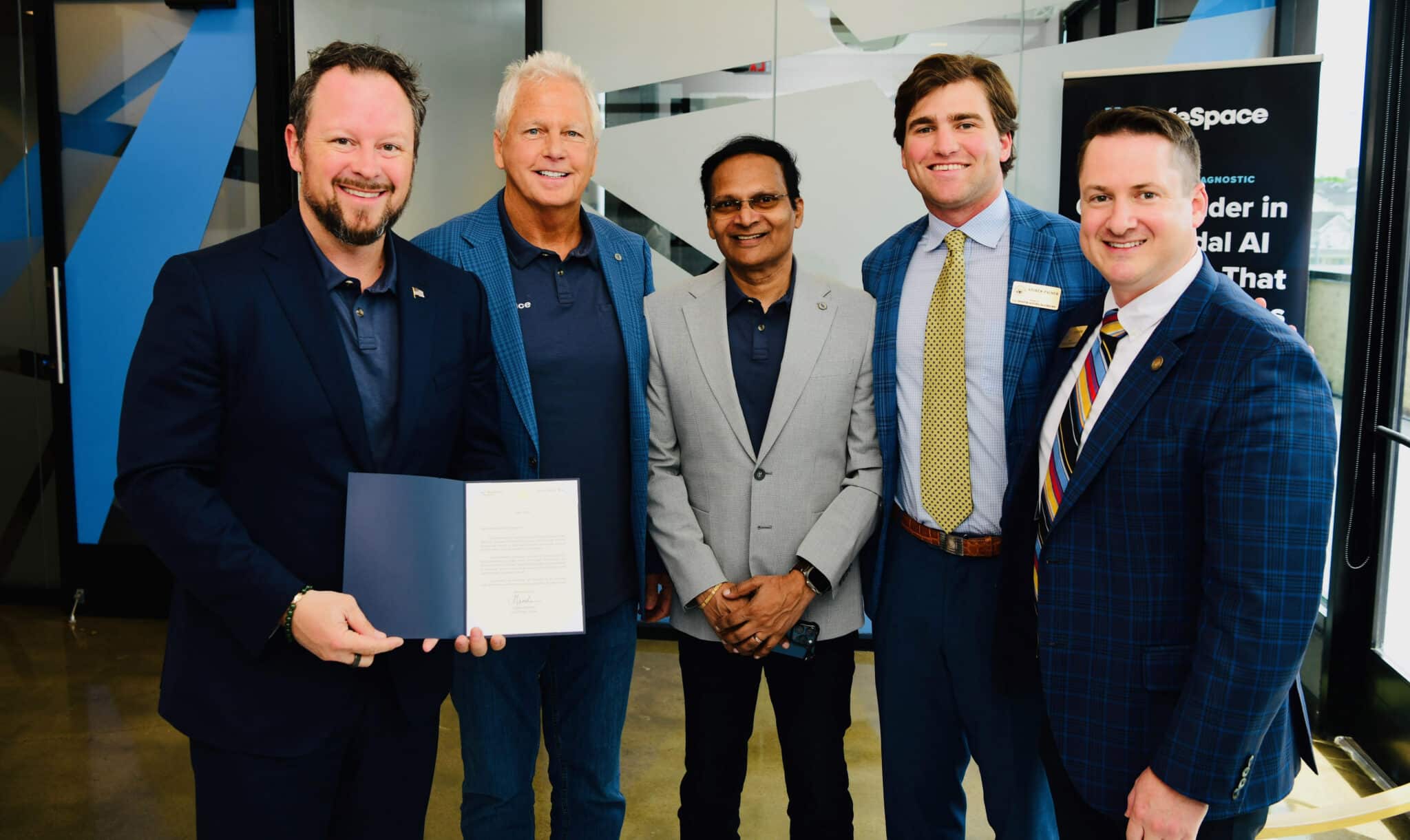 Five men stand indoors, with one holding an open document. They pose together, dressed in business or business-casual attire, smiling at the camera.