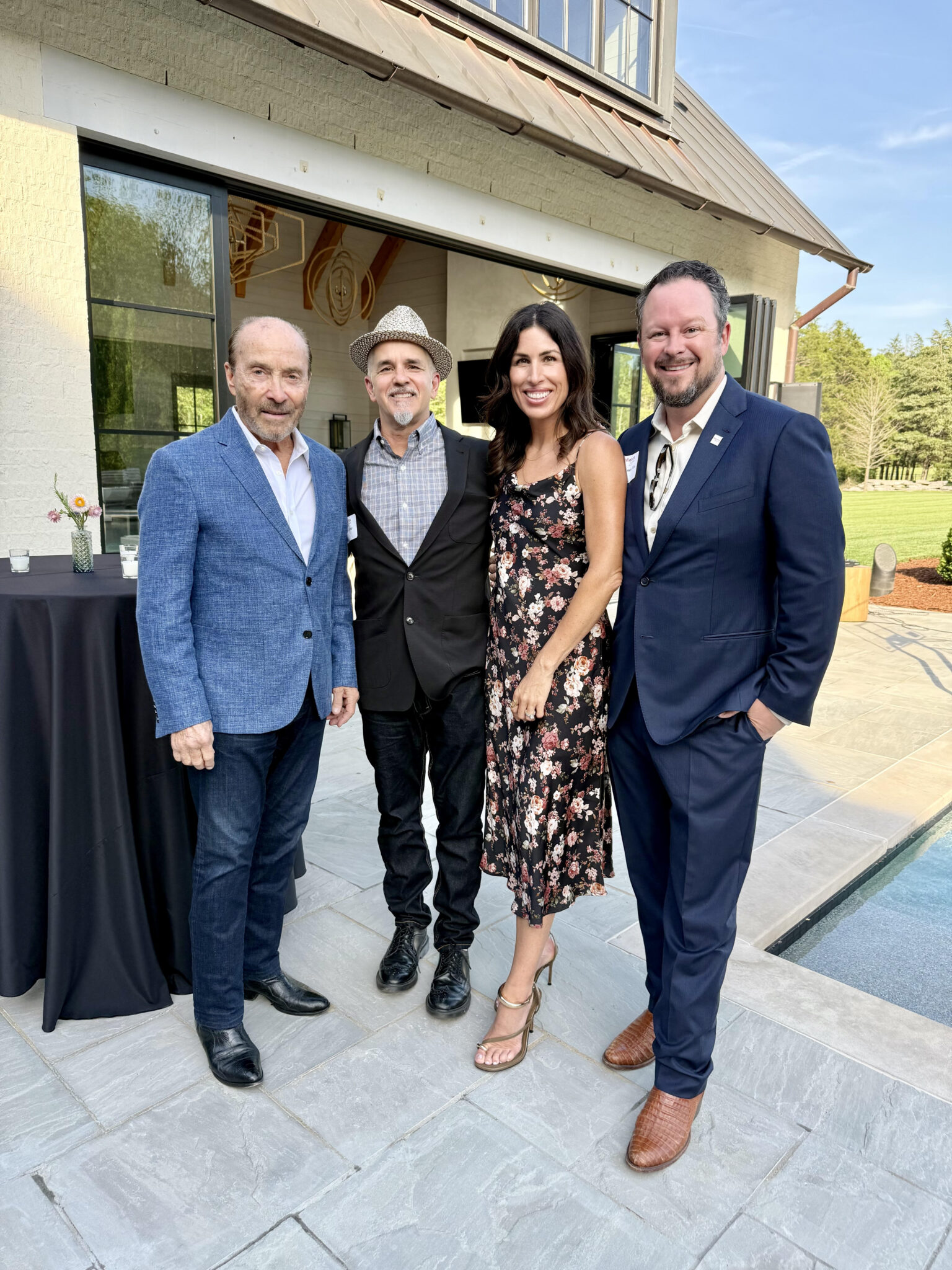 Four adults stand together outdoors on a stone patio near a pool, dressed in semi-formal attire, posing for a group photo at a social event.