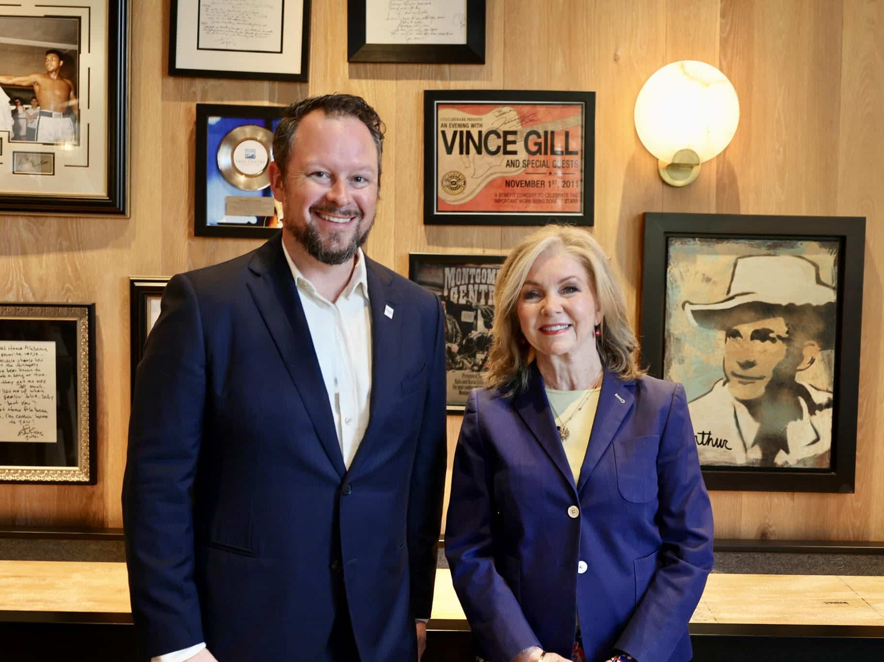 A man and a woman in blue suits stand side by side in front of a wood-paneled wall decorated with framed photos and memorabilia.