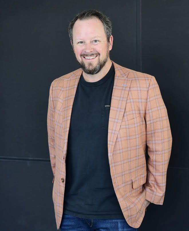 A man with a beard wearing a peach plaid blazer over a black t-shirt and jeans stands smiling in front of a plain dark background.