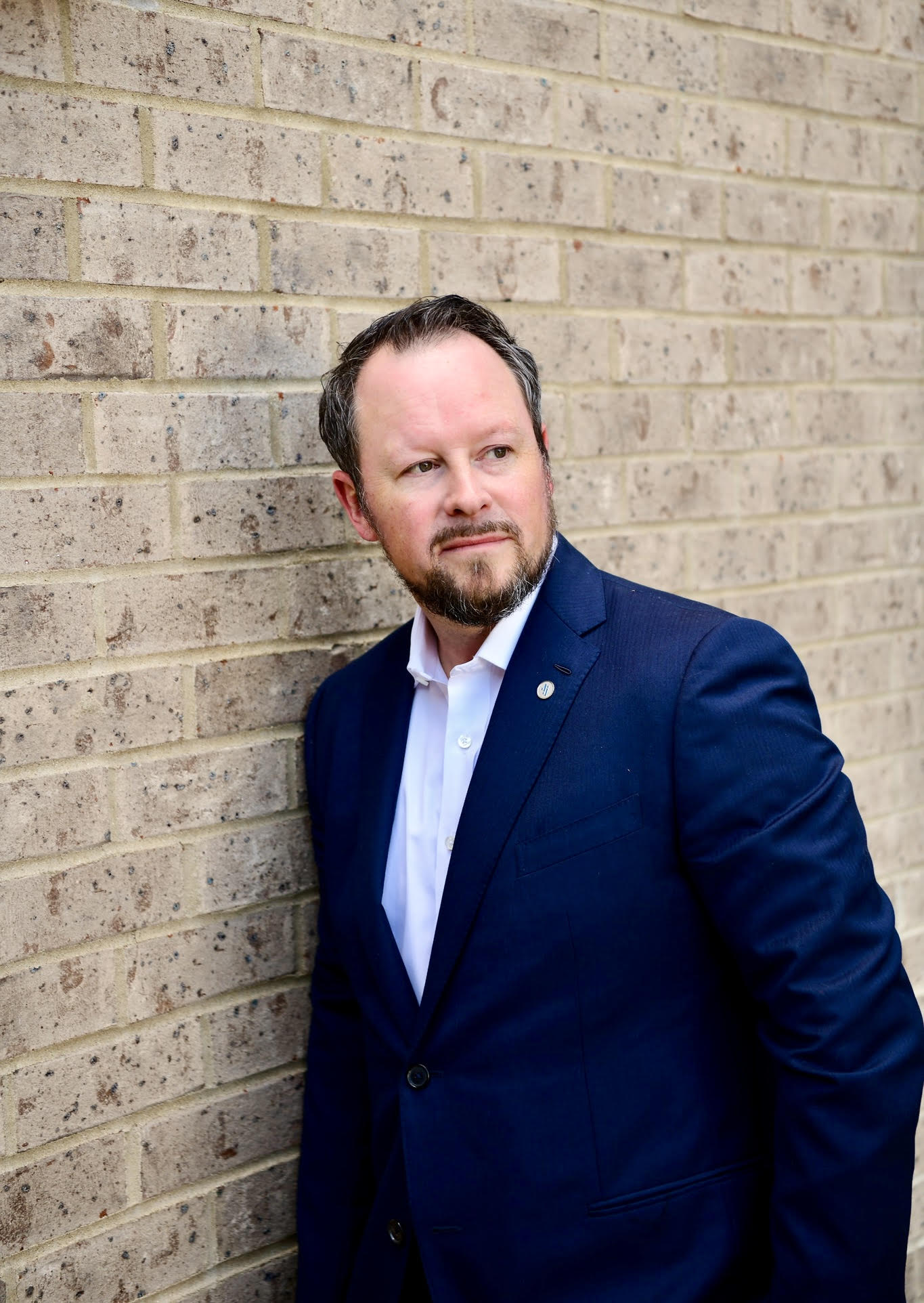 Man in a blue suit and white shirt stands against a brick wall, looking to the side with a neutral expression.