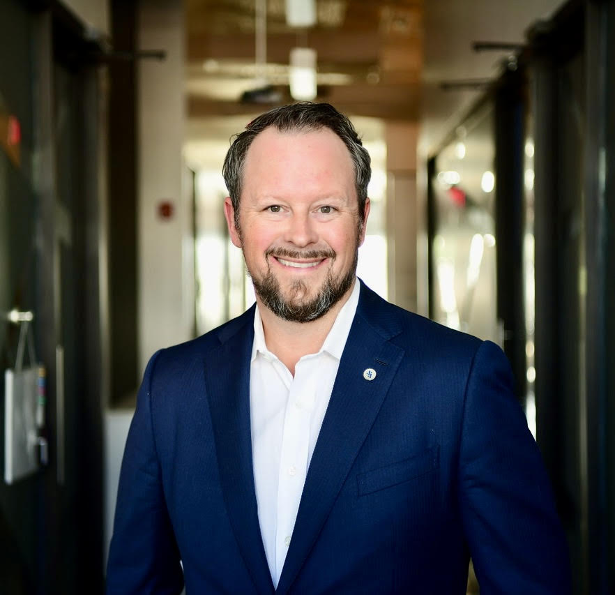 A man in a blue suit and white shirt stands in a well-lit hallway, smiling at the camera.